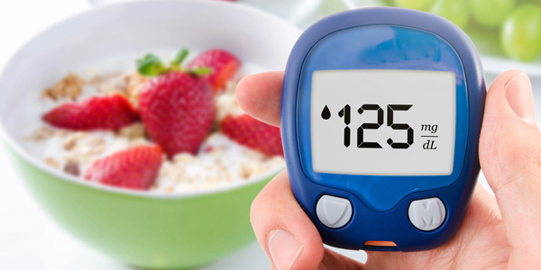 Eine Hand hält ein Blutzuckermessgerät mit der Anzeige ‚125 mg/dL‘; im Hintergrund steht eine Schale mit Müsli und Erdbeeren.